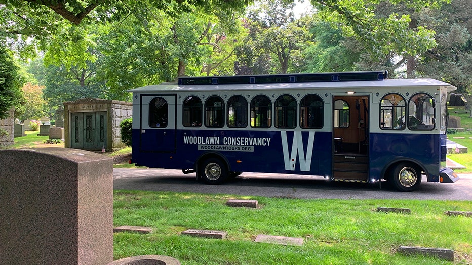 A blue and white bus designed to look like a classic trolley parked along a path in a scenic cemetery
