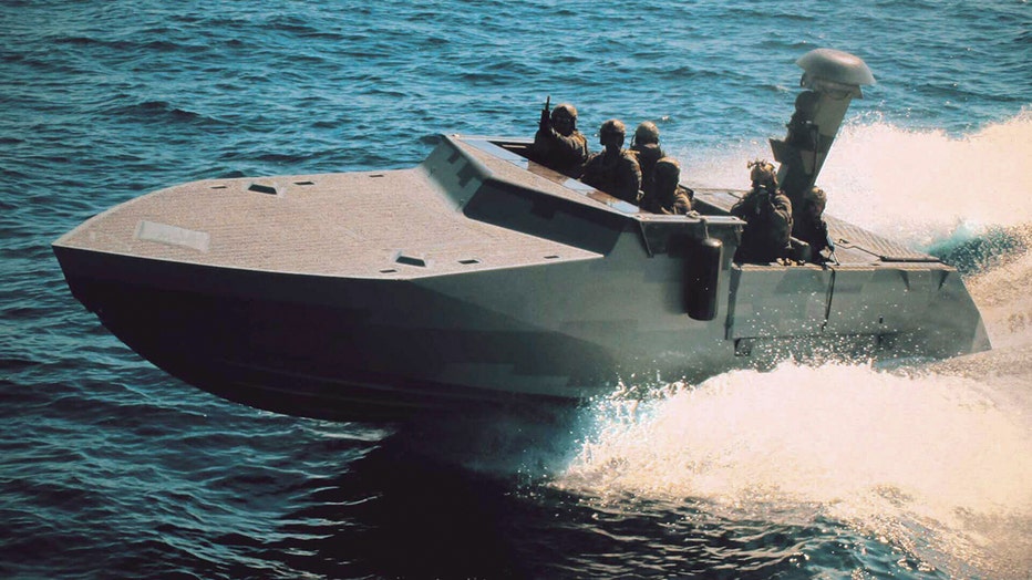 A slate gray navy vessel cuts through the water; several sailors are on board