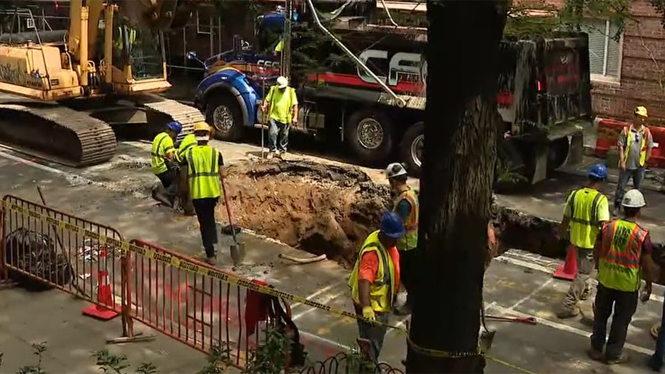 Several workers in hard hats and yellow safety vests work around a large hole in the surface of a tree-lined roadway in Manhattan