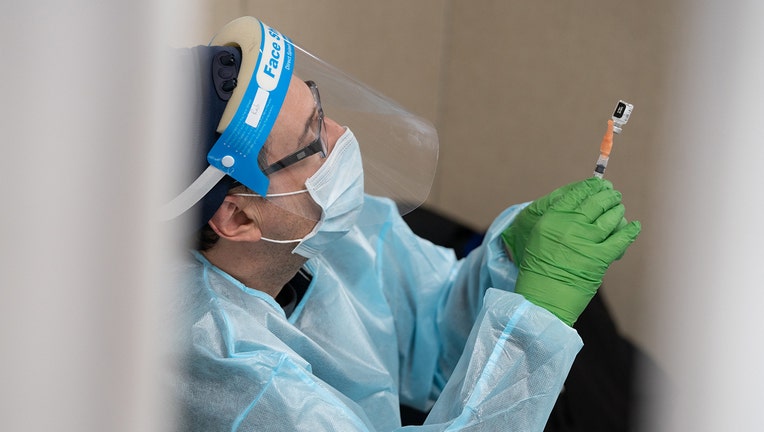 A medical worker wearing green gloves, a light blue mask and gown, and a clear face shield holds up a syringe