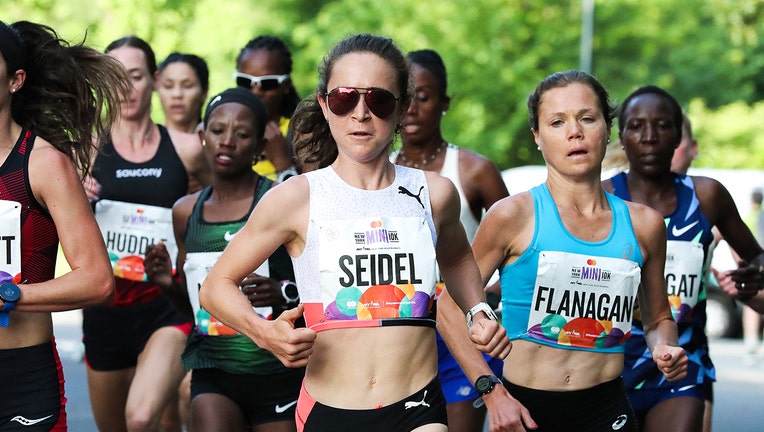 Several women running a race in Central Park; athlete at center of photo wears red aviator sunglasses, white tank top, dark briefs and a big with her name 'Seidel'