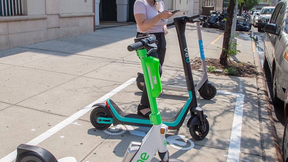 woman wearing helmet and mask looks at her mobile phone as she stands next to an electric scooter on the sidewalk; two more scooters are near her
