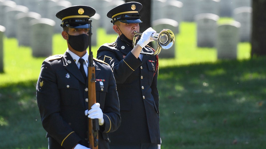 An Honor Guard soldier plays a bugle as another soldier, holding a rifle, stands at attention in a cemetery