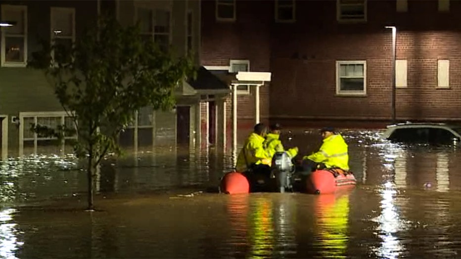 3 people navigate flooded residential streets in an inflatable boat; the crew are wearing fluorescent gear