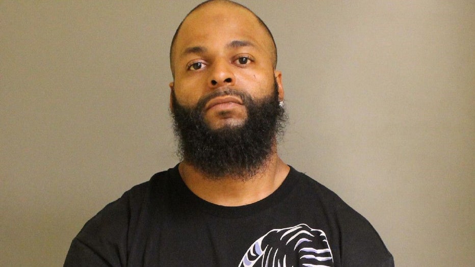 Head-and-shoulders booking photo of a man with a beard and short-cropped hair, wearing an earring and a black shirt