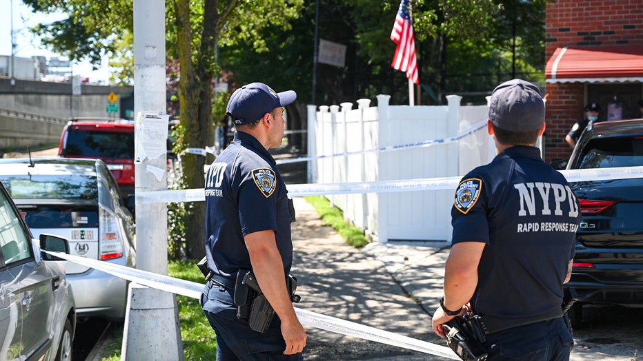 2 officers wearing blue polo shirts and ballcaps stand outside a residence in Queens