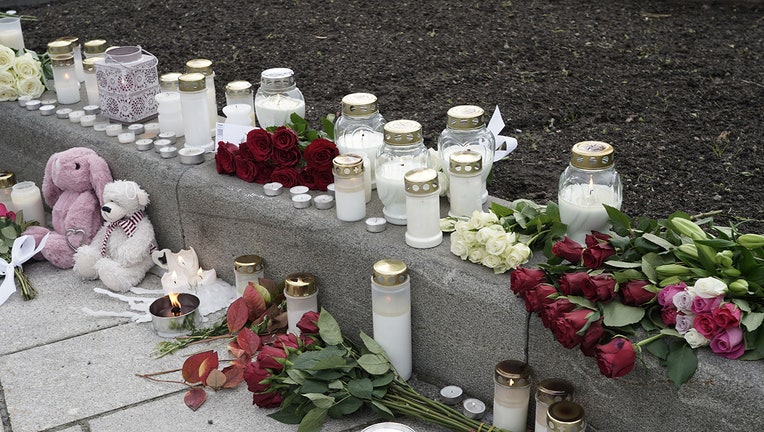 Several bouquets of roses, several candles, and two stuffed animals are left on a sidewalk