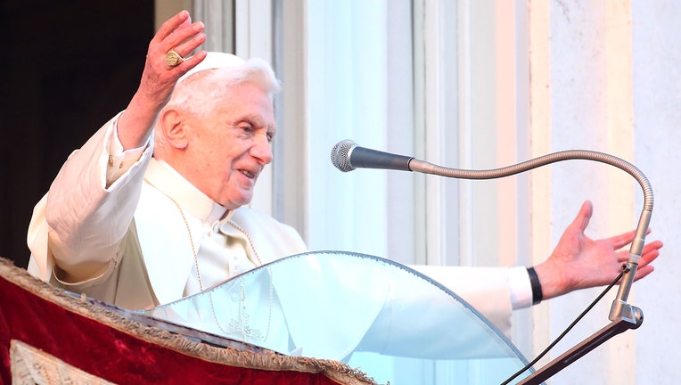 Pope Benedict in white vestments stands behind a lectern and microphone and waves