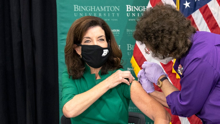 Governor wearing a black mask and green desk receives a booster shot in her left arm; a medical worker wearing a white mask and purple shirt administers the shot