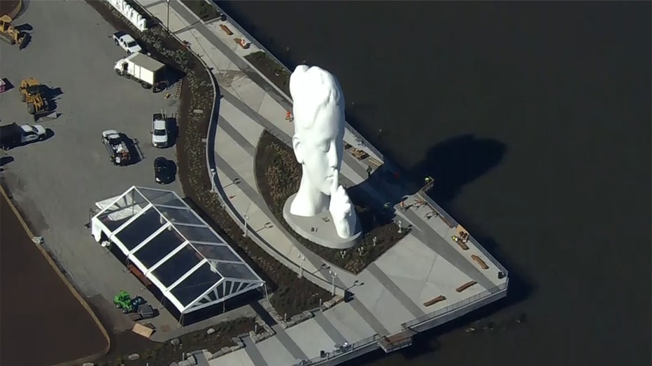 Aerial photo of a white 80-foot sculpture on a pier in the Hudson River; the sculpture depicts a face of a young woman holding a finger to her lips; several cars and trucks are near the sculpture