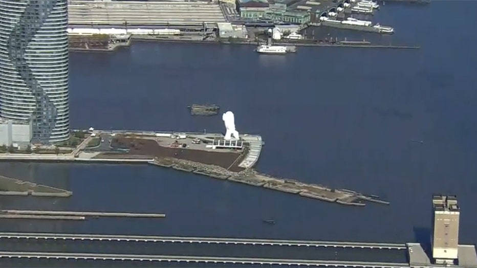 A long distance aerial photo of a white 80-foot sculpture on a pier in the Hudson River; the sculpture depicts a face of a young woman holding a finger to her lips; the image shows the pier and some buildings and structures nearby