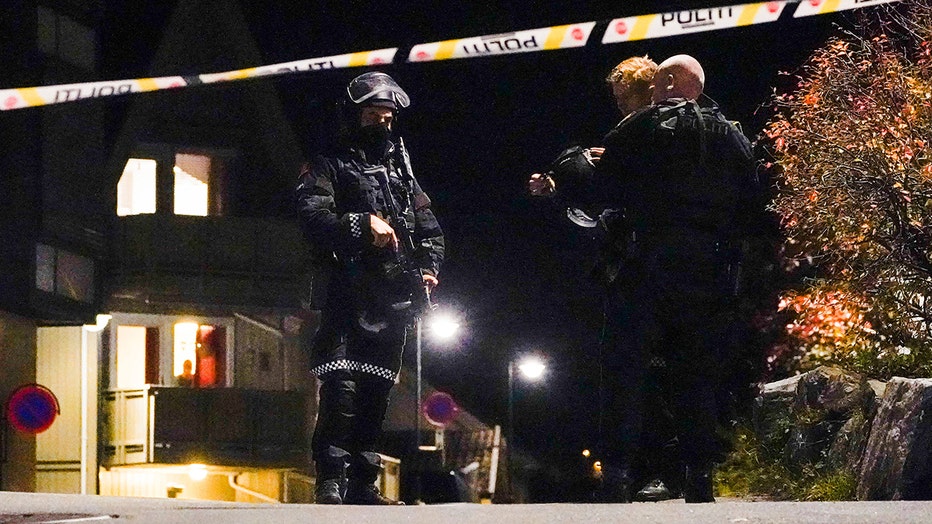 Nighttime scene showing a police officer armed with a long gun and wearing a helmet and mask and two other police officers standing behind police tape