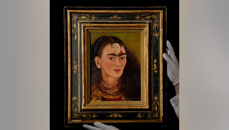 Painting by Frida Kahlo in a black and gold frame, held by a person's hands in white gloves; the painting is a self-portrait of the artist