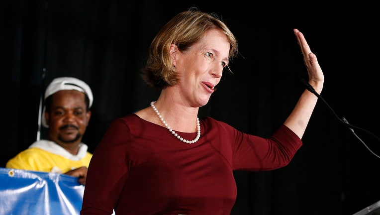 Woman waves, she is wearing a red shirt and white pearl necklace; a man stands in the background