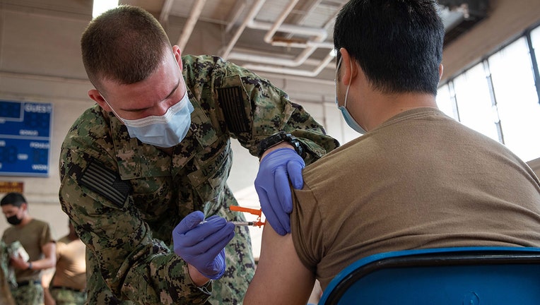 A sailor wearing a light blue mask and camo fatigues gives an injection to a sailor wearing a brown shirt