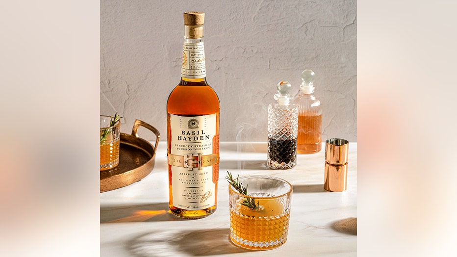 A cocktail garnished with a toasted rosemary sprig in a rocks and a bottle of bourbon