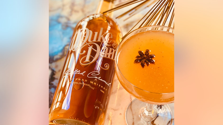 A caramel-colored cocktail garnished with star anise in a coupe glass and a whiskey bottle