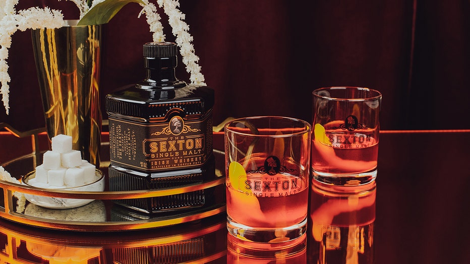 2 cocktails in rocks glasses with lemon peels, a octagonal-shaped whiskey bottle and a small bowl of sugar cubes
