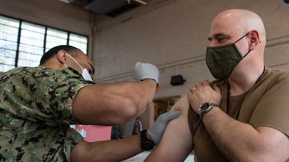 A sailor wearing a mask, gloves and camo fatigues gives an injection to a sailor wearing a brown shirt and green mask