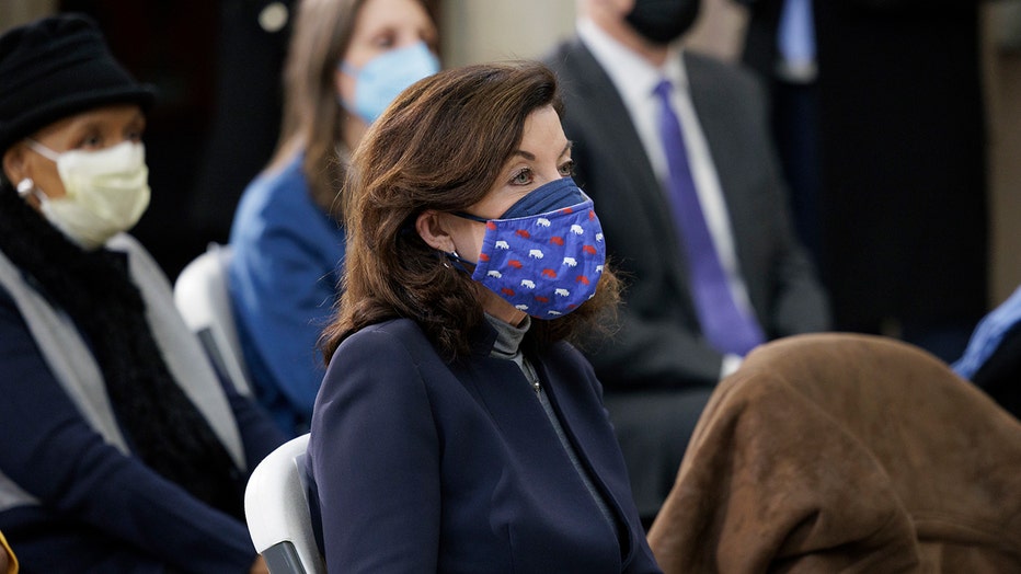 Gov Kathy Hochul sits among other people; she wears a blue suit, gray shirt, and a blue mask