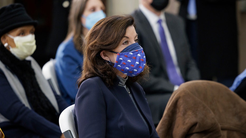 Gov Kathy Hochul sits among other people; she wears a blue suit, gray shirt, and a blue mask