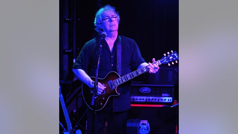 Musician Ian McDonald behind a mic and playing a guitar; he has white hair, dark shirt and glasses