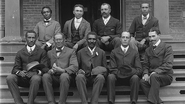 George Washington Carver, full-length portrait, seated on steps, facing front, with staff; 9 people are in this black-and-white image