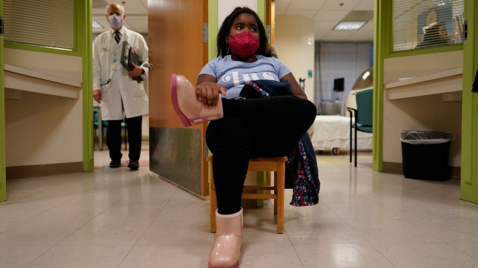 young girl in a red mask, blue shirt, dark pants and pink boots sits outside exam rooms in a hospital