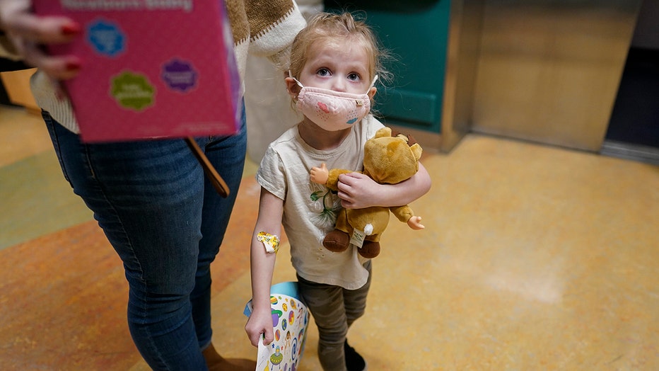 young girl wears a mask and holds a stuffed toy