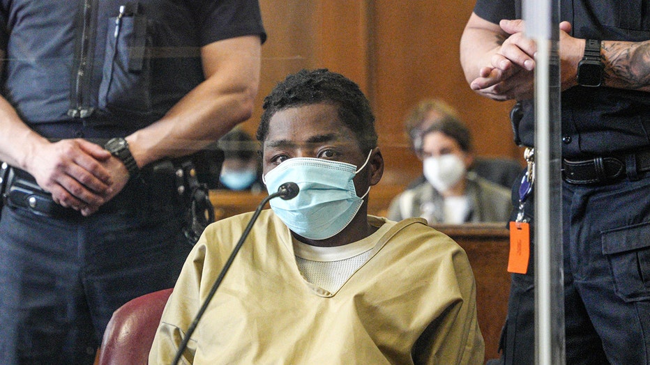 Man wearing light blue mask and tan jumpsuit sits in a courtroom