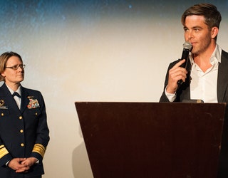 Actor Chris Pine speaks into a microphone and stands behind a lectern as Adm. Fagan, wearing a dark blue uniform stands nearby and watches