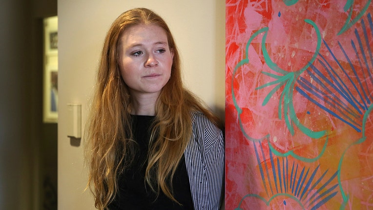 A woman with long red hair and wearing a striped shirt over a black top poses next to a colorful wall