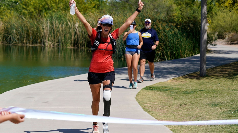 Runner raises her arms to celebrate finishing a marathon near a lake; she wears black shorts, a red t-shirt, sunglasses and a cap; her left leg is a "blade runner-style" prosthetic