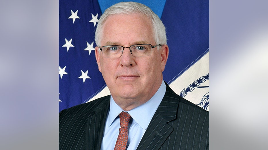 Head-and-shoulders photo of John Miller; he is wearing a gray suit jacket, light blue shirt, patterned tie, and glasses; he has white hair; US and NYC flags hang behind him