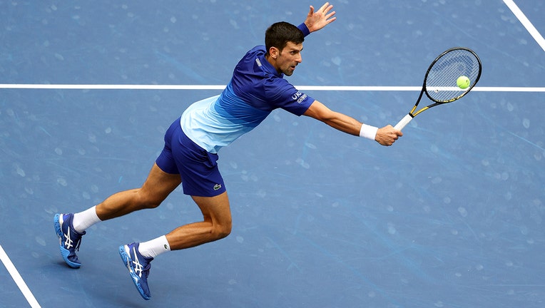Tennis player Novak Djokovic lunging to hit the ball on a blue tennis court; he wears a blue-and-white kit
