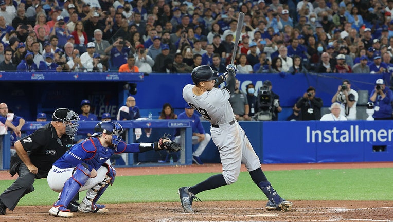 MLB player Aaron Judge swinging as he hits a home run; he wears a gray road uniform; behind him are a catcher and umpire; in the background are fans in the stands