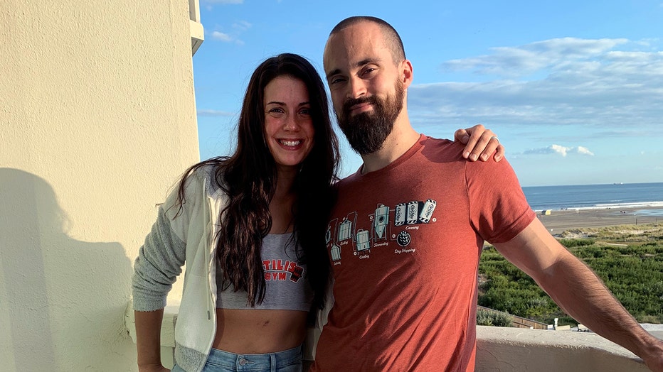 Woman and man standing on a balcony; they smile at camera; blue sky and a beach are seen in background