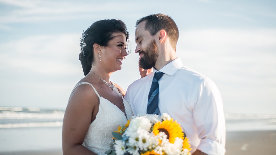 Wedding photo of a woman and man on the beach; they face each other; she wears a sleeveless white gown and holds a flower bouquet; he wears a white shirt, blue tie, and khaki pants