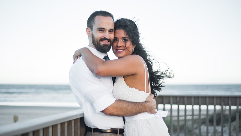 Wedding photo of a woman and man stand at a railing with beach behind them; they smile at the camera; she wears a sleeveless white gown; he wears a white shirt, blue tie, and khaki pants