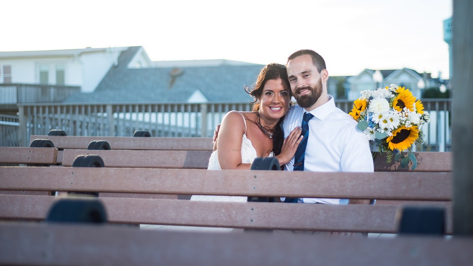 Wedding photo of a woman and man sitting on a bench on a boardwalk; they smile at the camera; she wears a sleeveless white gown and holds a flower bouquet; he wears a white shirt, blue tie, and khaki pants