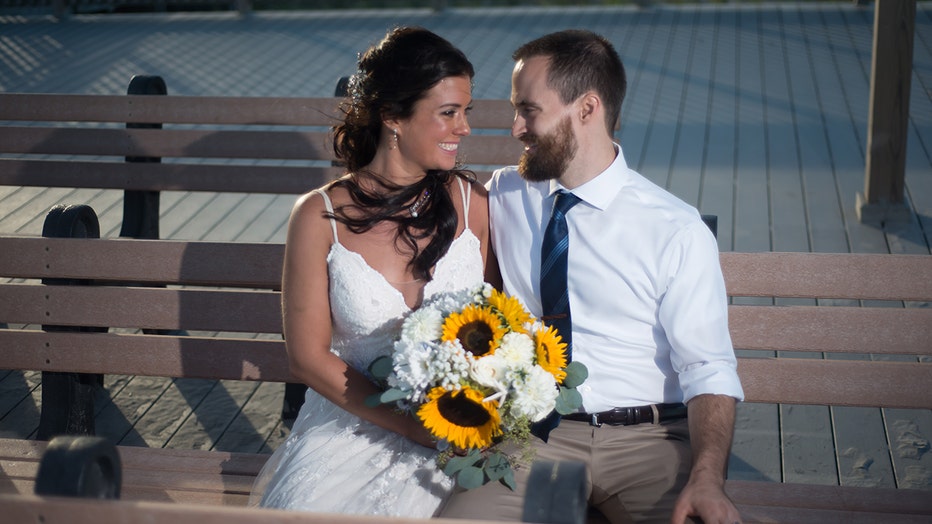 Wedding photo of a woman and man sitting on a bench on a boardwalk; they smile each other; she wears a sleeveless white gown and holds a flower bouquet; he wears a white shirt, blue tie, and khaki pants