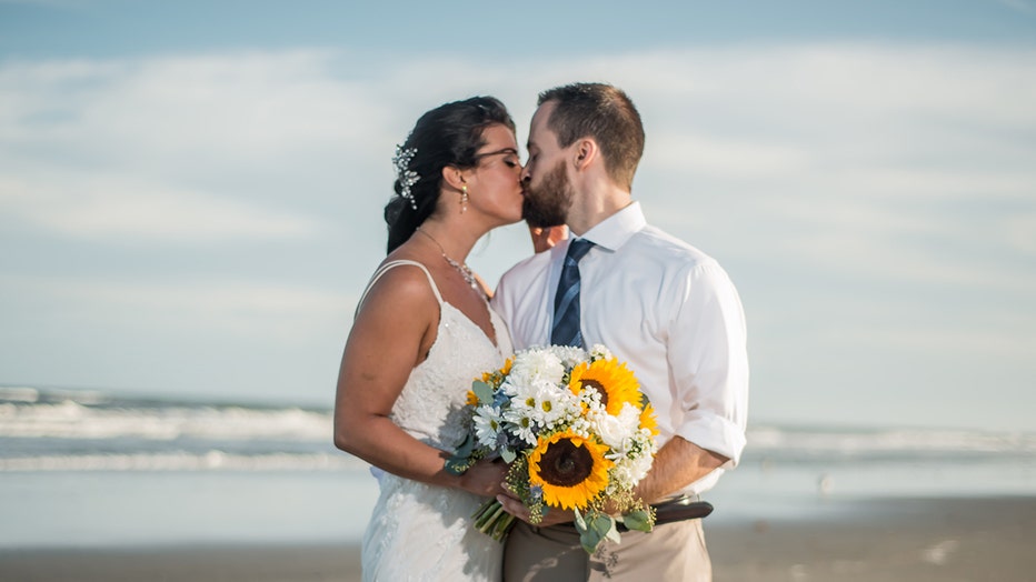 Wedding photo of a woman and man on the beach; they kiss; she wears a sleeveless white gown and holds a flower bouquet; he wears a white shirt, blue tie, and khaki pants