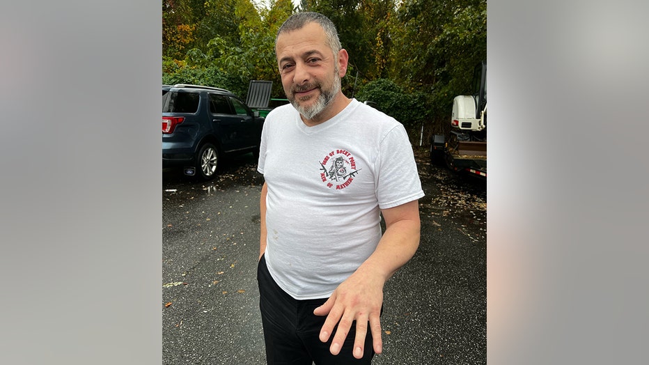 A bearded man wearing a white T-shirt and black pants holds out his hand to show cuts on his fingers