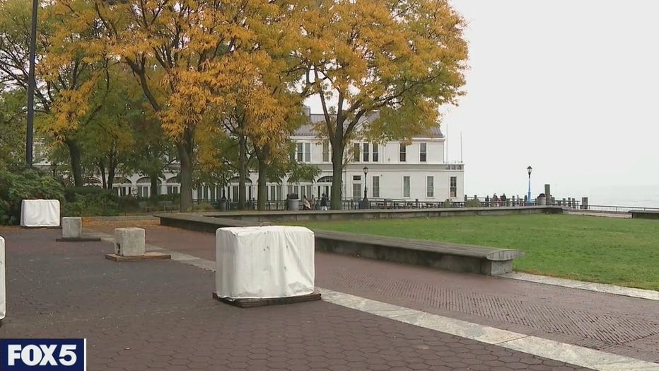 Trees, grass, benches and walkways in a park; a building and water are in the background