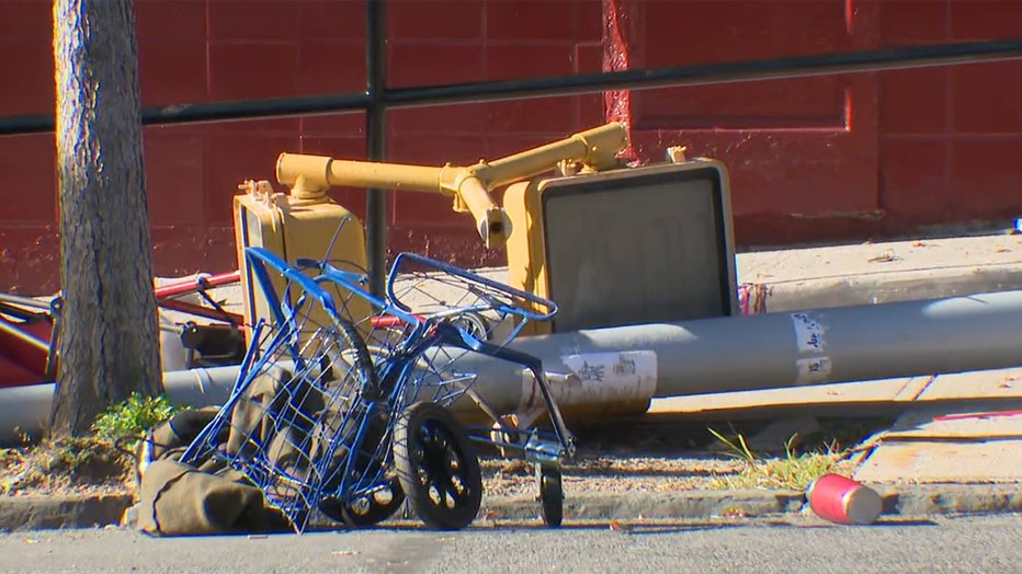 A grocery cart lies crumpled on the edge of a road and a crosswalk traffic signal pole is toppled