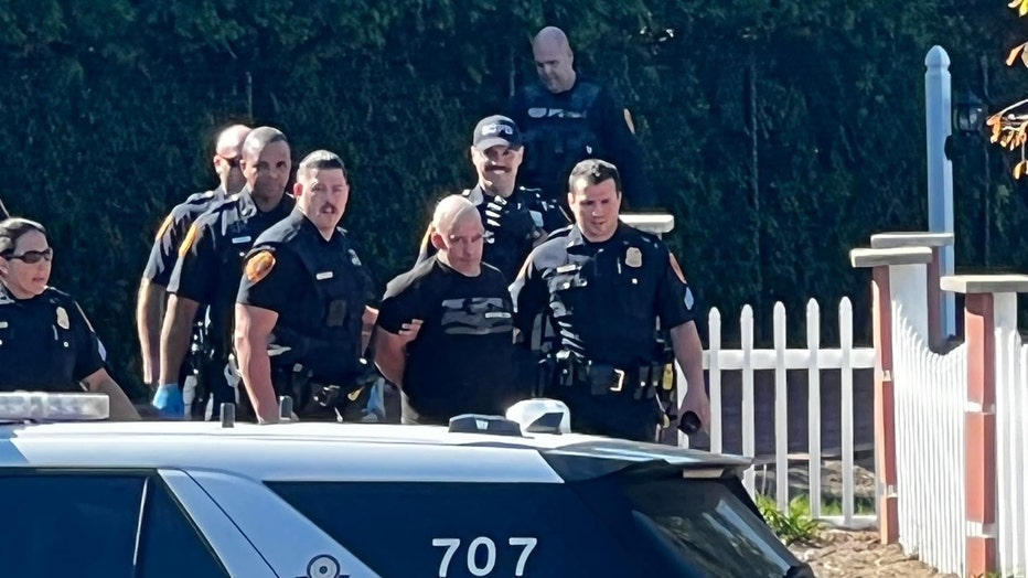 Seven police officers escort a handcuffed man from the woods to a police car