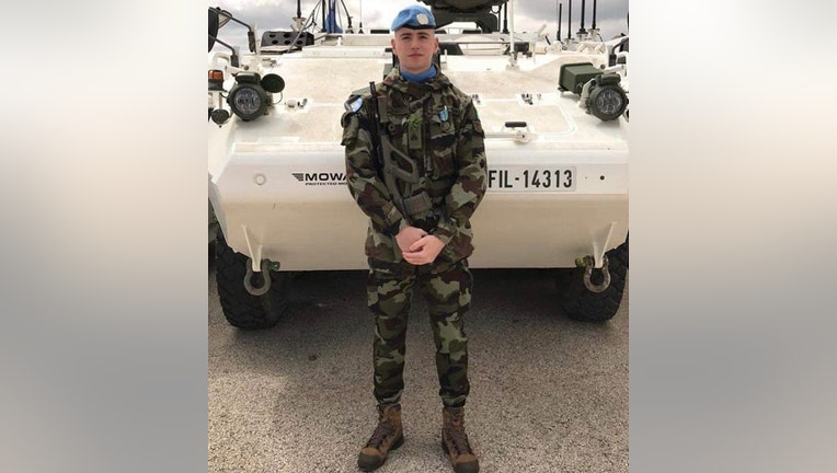 U.N. peacekeeper wearing a camouflage uniform and a blue beret stands in front of an armored vehicle
