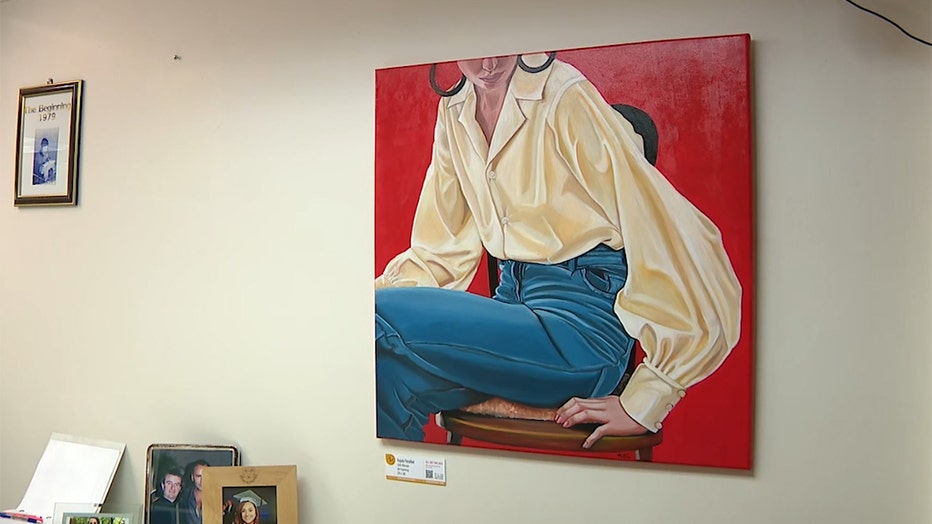 An oil painting on a wall; the painting shows a figure in a blouse and trousers sitting on a chair