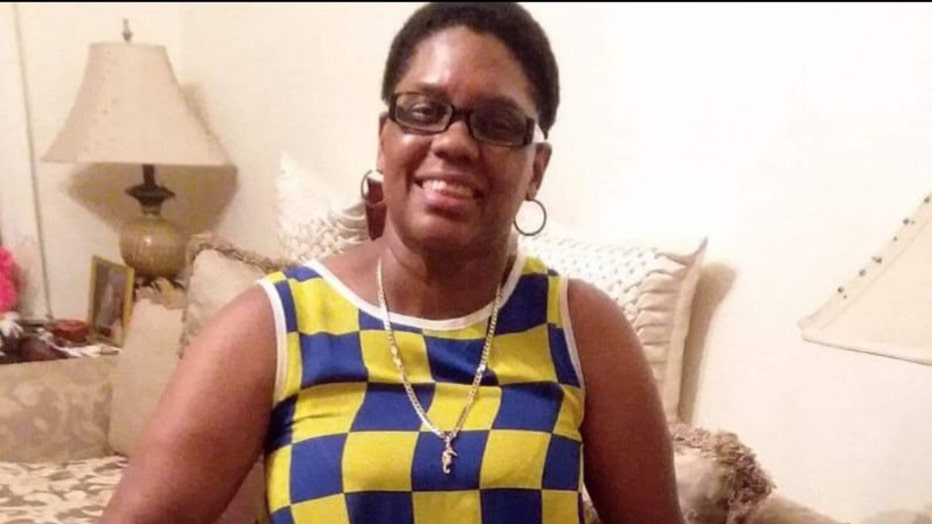 Woman smiling; she has short hair; she wears a blue and yellow checkered tank top, hoop earrings, a long necklace, and glasses