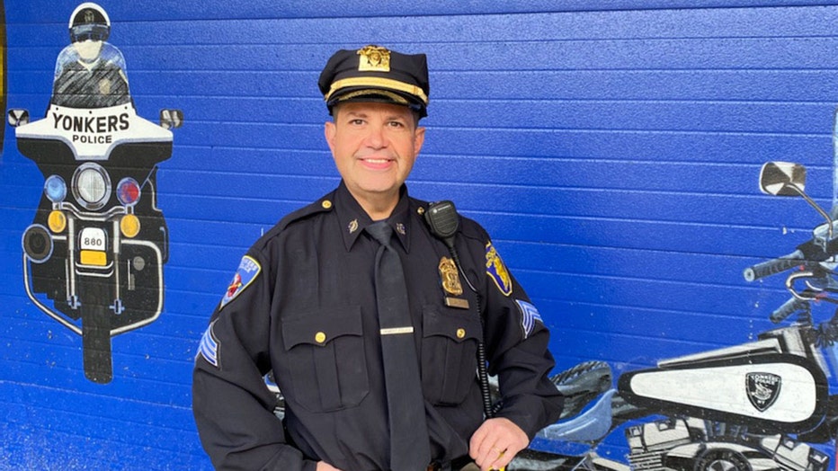 A police sergeant poses in uniform in front of a blue wall with a mural